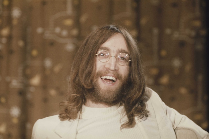 A man smiles widely. He wears white turtle-neck and thin-rimmed round glasses. He has shoulder-length hair, moustache and beard.