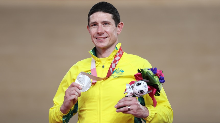 An Australian male cyclist smiles as he holds his Tokyo Paralympic medal on the podium.