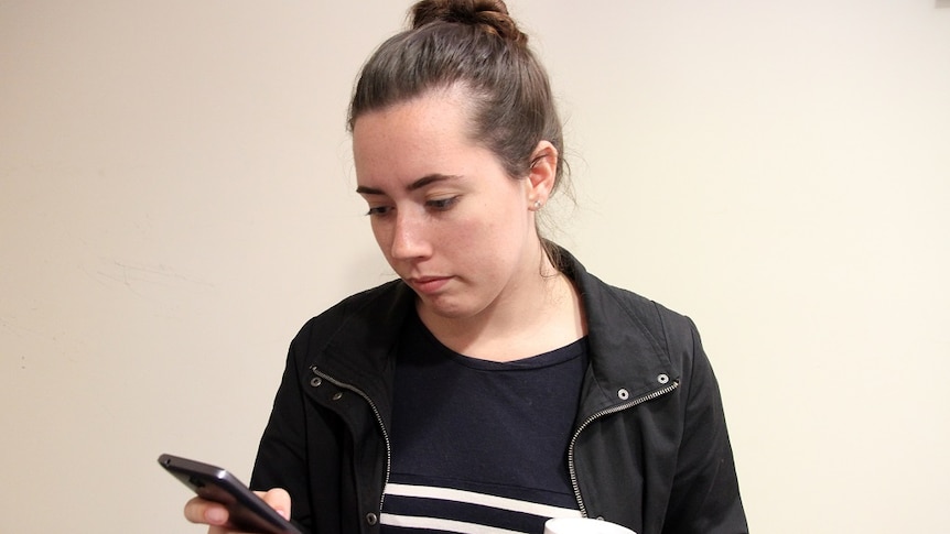 A 20-year-old woman is sitting on a chair and is looking at her phone with a take-away coffee in her hand.