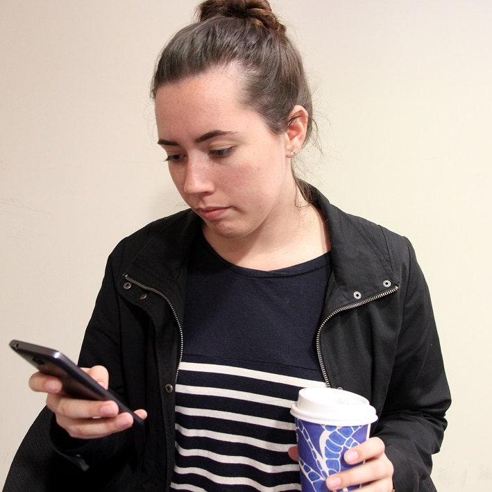 A 20-year-old woman is sitting on a chair and is looking at her phone with a take-away coffee in her hand.