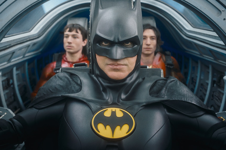 Two versions of Barry, a white man with dark hair, sit in the backset of a jet being flown by Batman in bat suit.
