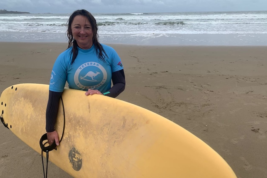 Woman stands on beach holding a surf board and smiling