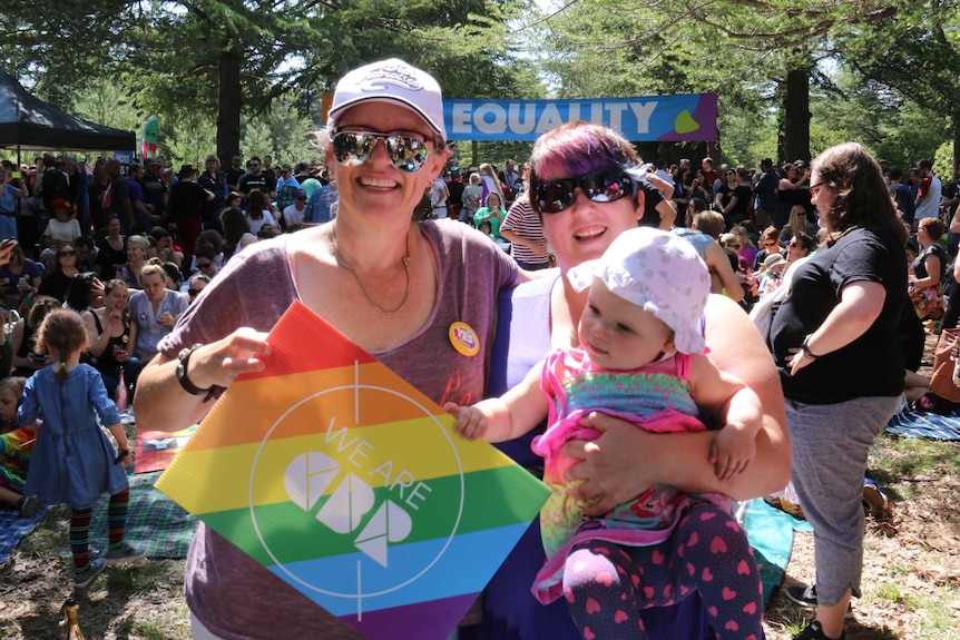 Two women, one holding a baby, smile in front of a crowd in a park.