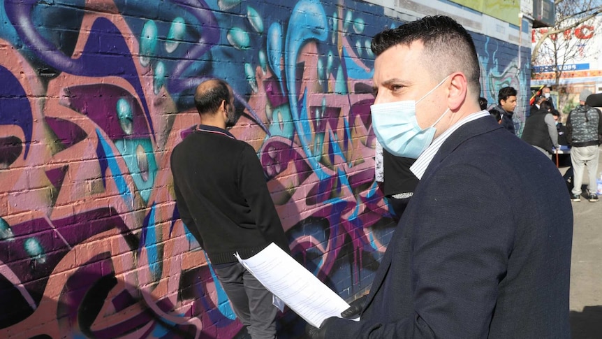 A man with a surgical mask stands in front of a colourful mural.