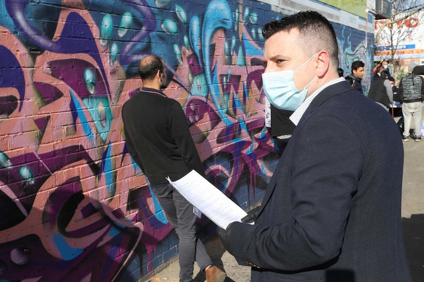 A man with a surgical mask stands in front of a colourful mural.