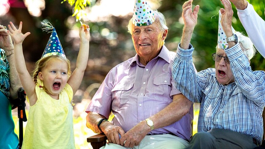 An elderly man in a party hat throws up his hands with a young girl on the ABC TV series Old People's Home For 4 Year Olds.