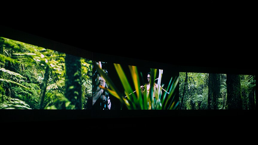 In dark room, two screens of lush forest scenes, border a screen of bare chested man holds fighting staffs over shoulders.