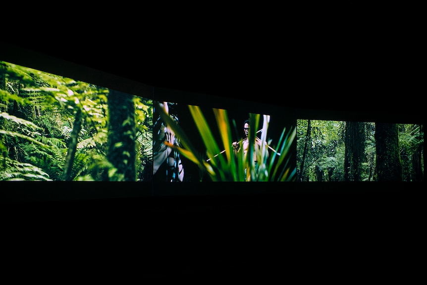 In dark room, two screens of lush forest scenes, border a screen of bare chested man holds fighting staffs over shoulders.