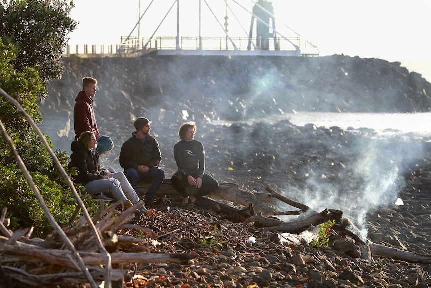 Five young people in wetsuits and warm clothes huddle by a fire on the rocks