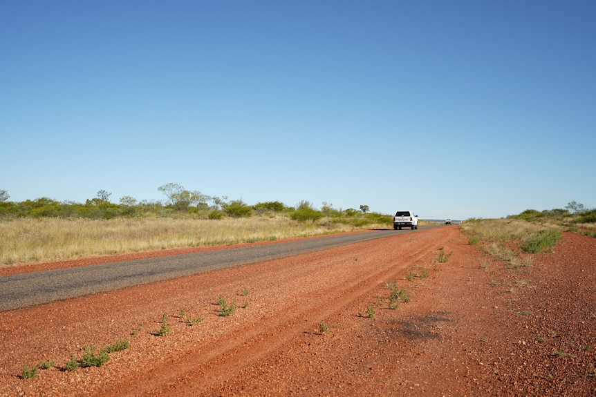 A car driving on a small, one lane sealed road in outback Queensland, blue skies, red dirt, scrub, green on the side.