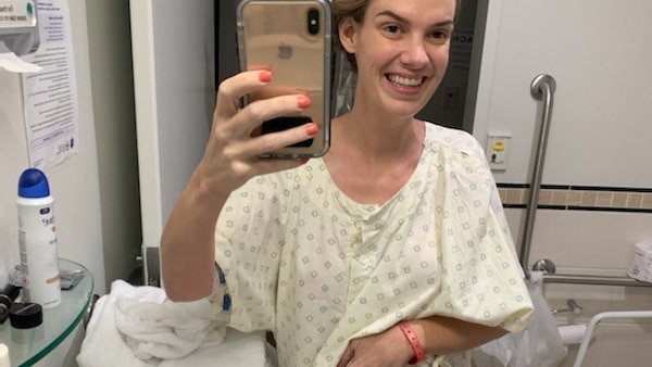 A woman takes a selfie in a hospital bathroom mirror, wearing a hospital gown, bag showing, smiling.