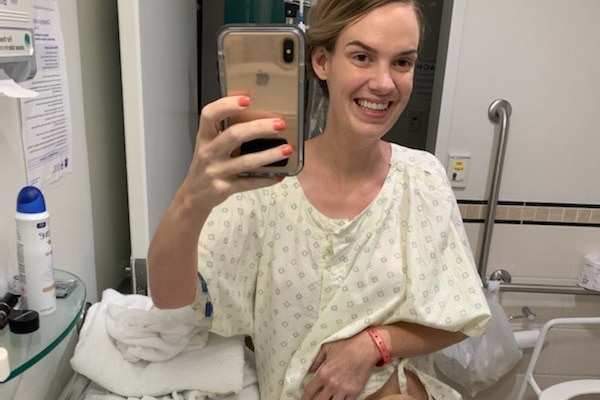 A woman takes a selfie in a hospital bathroom mirror, wearing a hospital gown, bag showing, smiling.