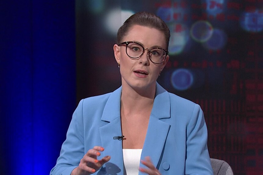 A woman wearing a light blue suit jacket and glasses speaking while moving her hands