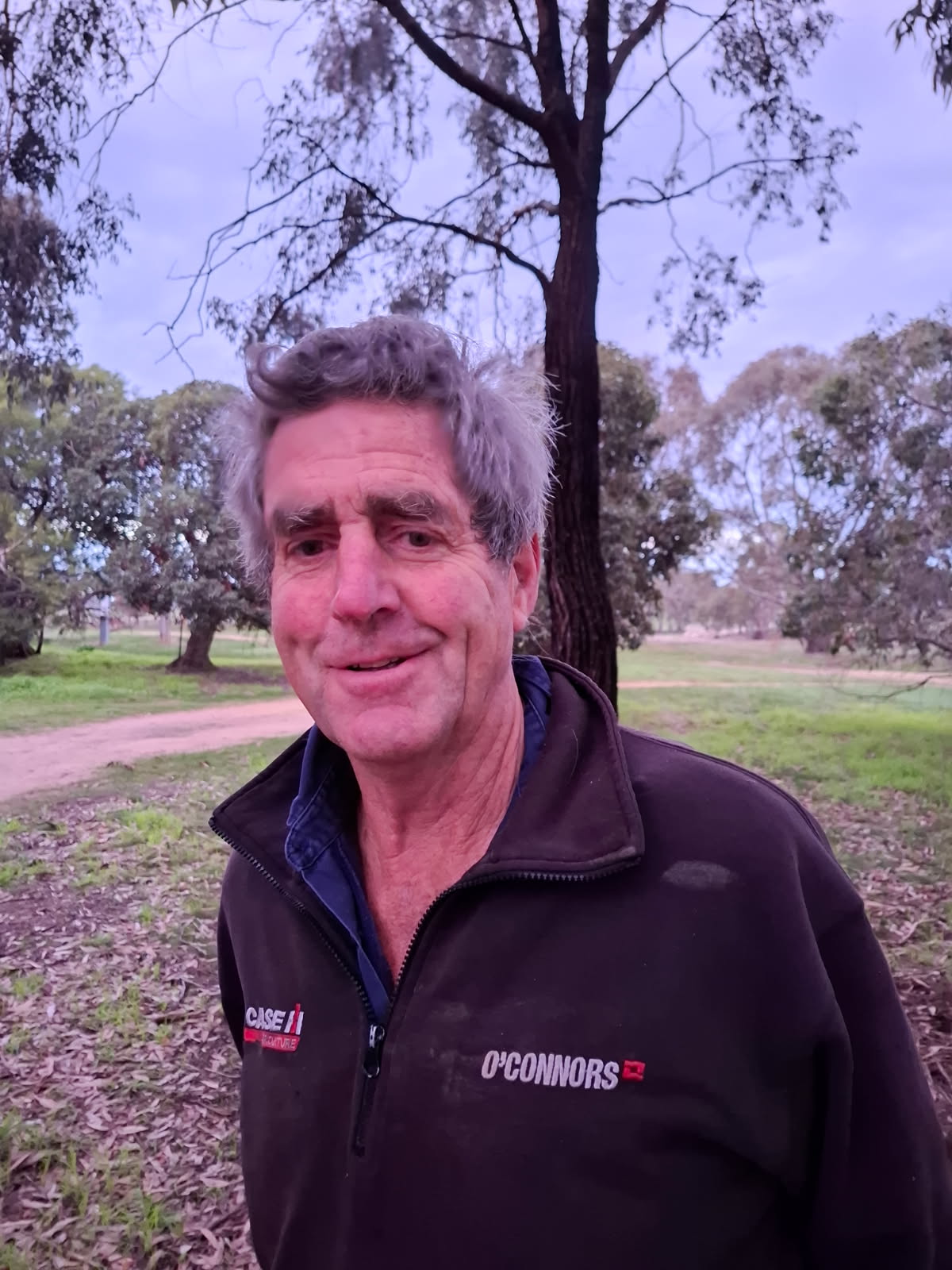 An older man with greying hair stands near a dirt driveway and some trees on a country property.