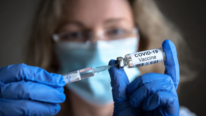 A woman in a mask and gloves uses a needle to pull liquid from a vaccine vial