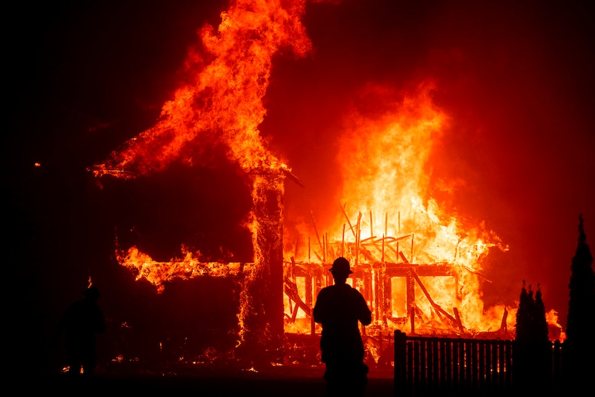 A person stands in front of a house in Paradise that is burning.
