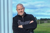 Portrait of Mark Pember leaning against a fence with arms folded outside 