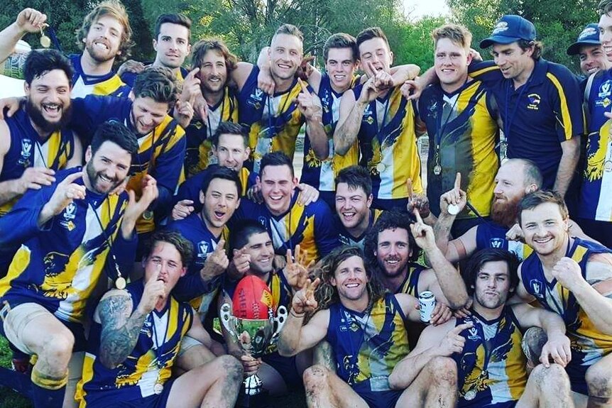 A group of delighted footballers pose around a premiership cup, celebrating a win.
