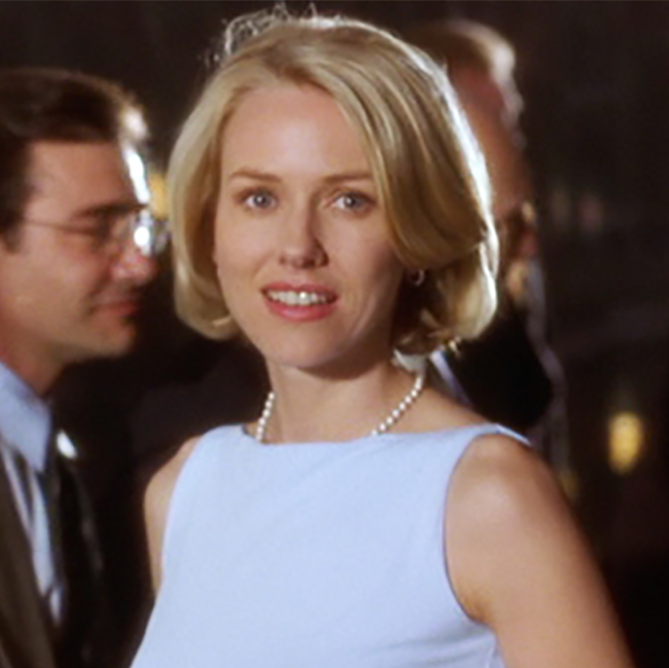 Actor Naomi Watts wearing a light blue dress and pearls, smiling at the camera while a group of people mingle in the background.
