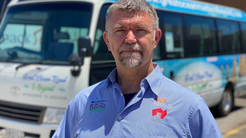 A man wearing a blue collared shirt stands with a grim expression in front of a tour bus