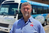 A man wearing a blue collared shirt stands with a grim expression in front of a tour bus