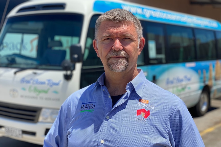 A man wearing a blue collared shirt stands with a grim expression in front of a tour bus