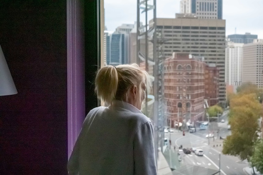 A blonde woman looks out a hotel room window.
