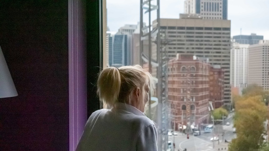 A blonde woman looks out a hotel room window.
