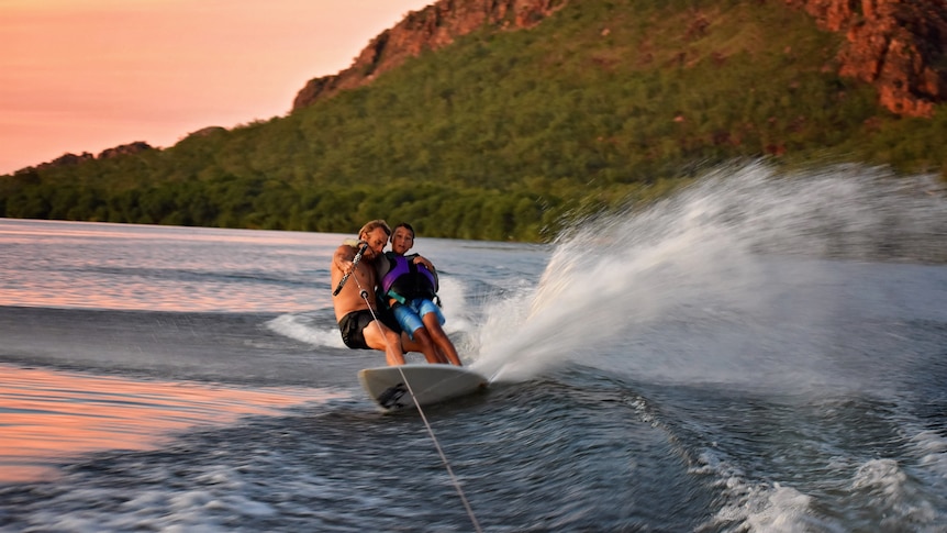 Man with younger boy on surfboard being towed behind boat, with hills in background.