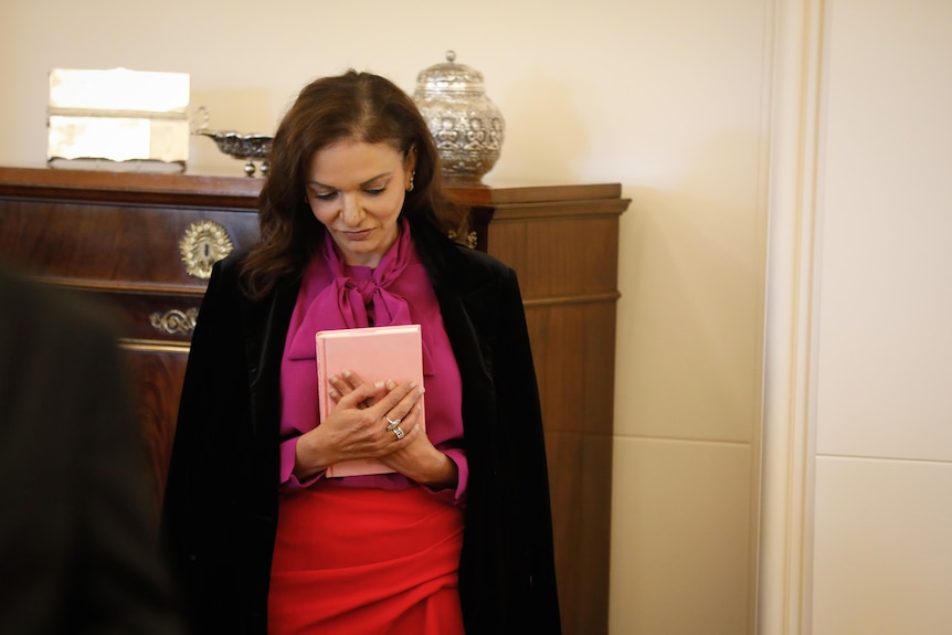 Aly holds a pink Koran to her chest and smiles as she looks down, while standing inside Government House.