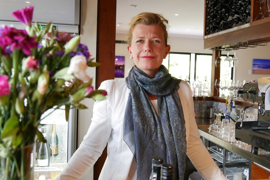 Woman wearing white jacket and grey scarf, with blonde hair pulled back, stands at restaurant counter and looks into the camera.