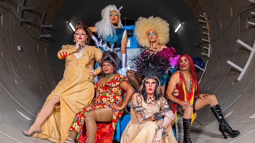Six Indigenous Drag Queens pose together in full drag looking glamorous and fierce.