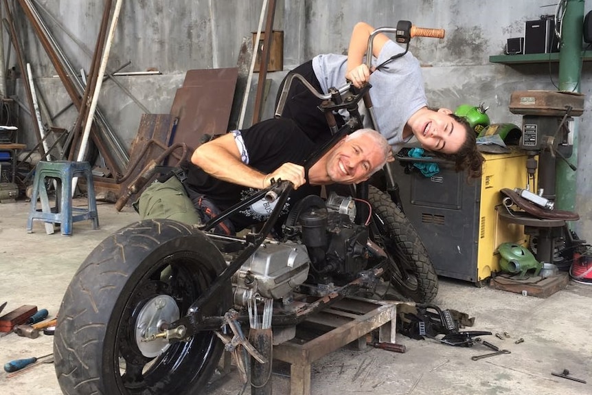 A smiling man and a girl leaning on a motorbike