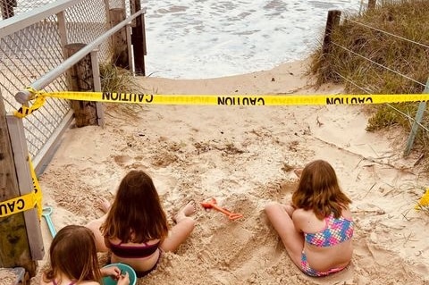 Children play in the sand behind caution tape, with waves crashing against eroded beach.