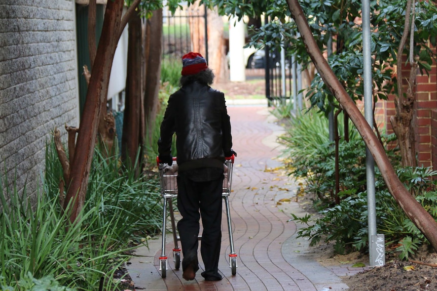 A homeless man walks through a public alley with his back to the camera, pushing a trolley.