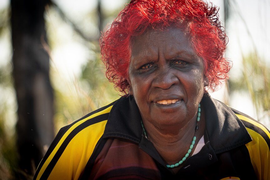 Woman with bright red died hair smiles at camera with trees in background