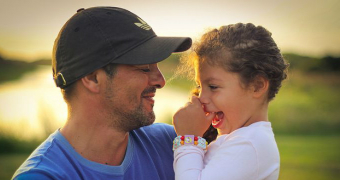 A dad wearing a black Adidas cap and blue shirt hugs his daughter wearing a pink shirt with her thumb in her mouth.