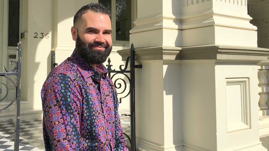 Young Indigenous man with beard, wearing a purple print shirt, stands in front of historic sandstone building.