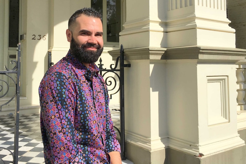 Young Indigenous man with beard, wearing a purple print shirt, stands in front of historic sandstone building.