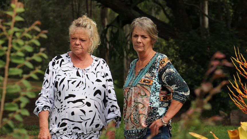 Two women stand together in a garden. One has a white shirt with animal print, another had a teal printed shirt.