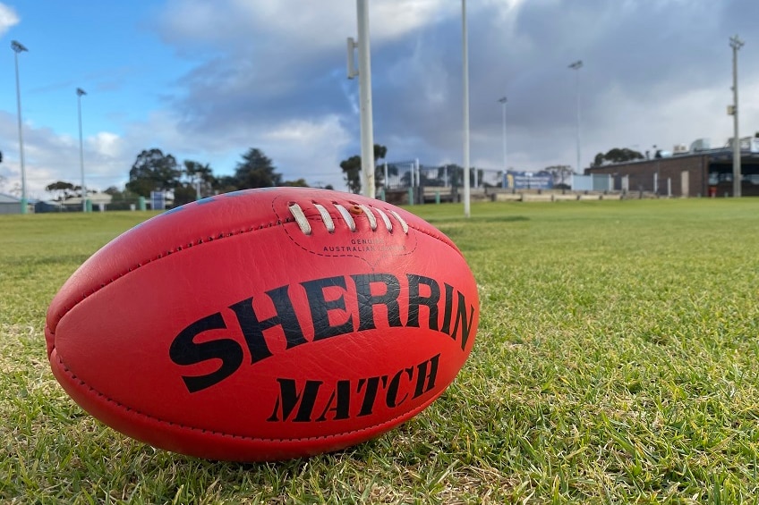 red sherrin football to sitting on green grass with football goal posts and football club in the background