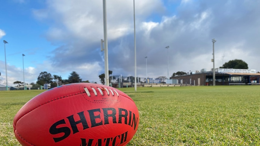 red sherrin football to sitting on green grass with football goal posts and football club in the background