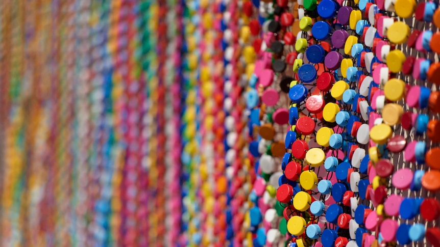 Many rows of bottle tops strung together to make up a blanket type artwork that's 12 metres long.