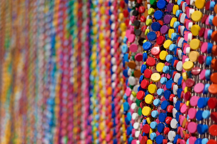 Many rows of bottle tops strung together to make up a blanket type artwork that's 12 metres long.
