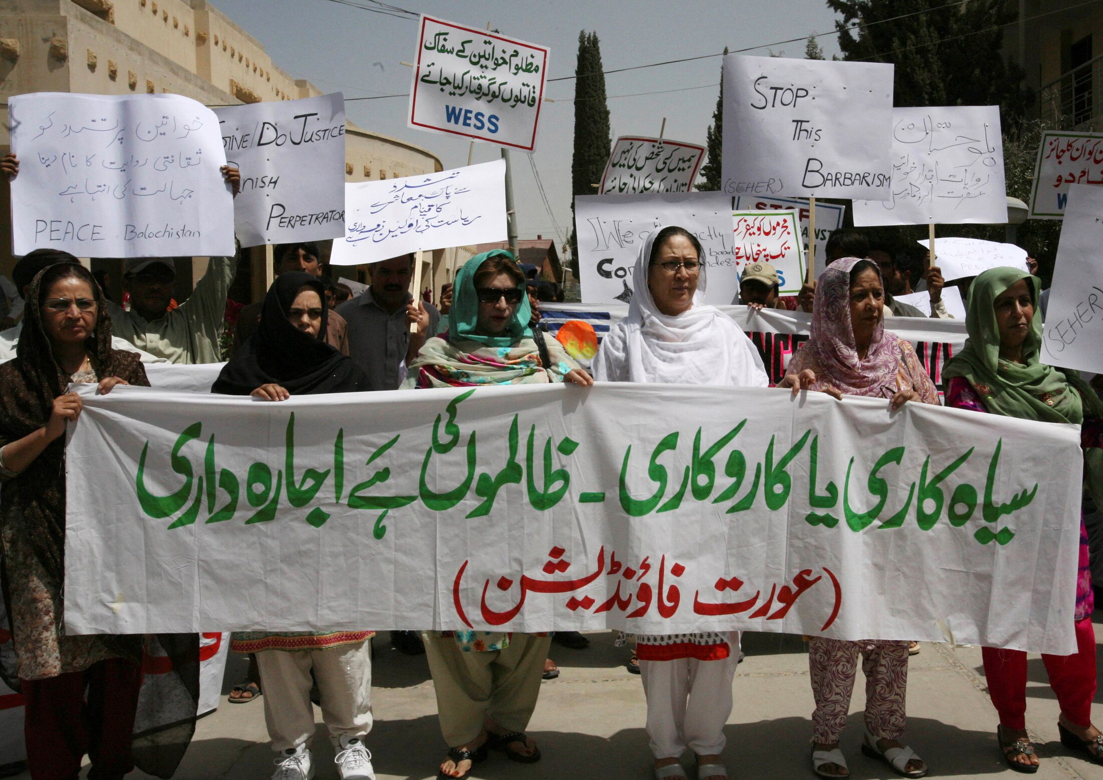 women wearing headscarves carry white cloth with green and red Arabic script. Behind are signs in English and Arabic.