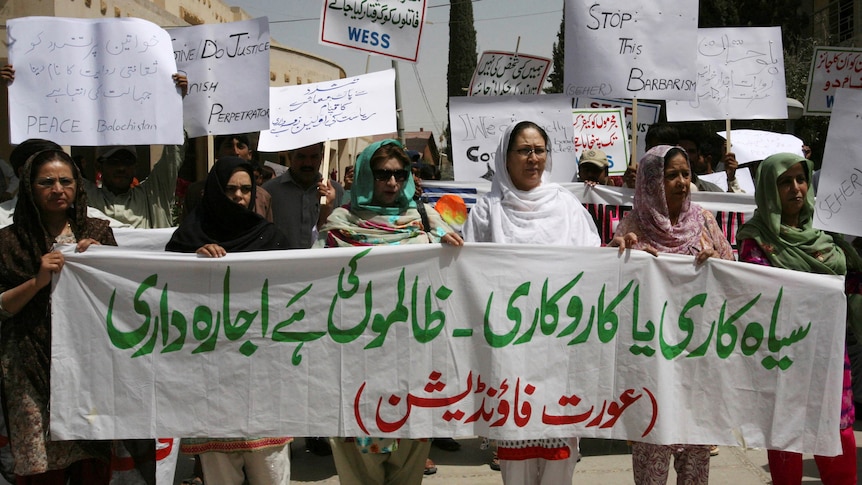 women wearing headscarves carry white cloth with green and red Arabic script. Behind are signs in English and Arabic.