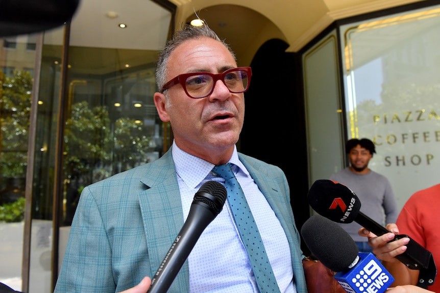 Andrew O'Keefe addresses the media wearing a suit and red glasses