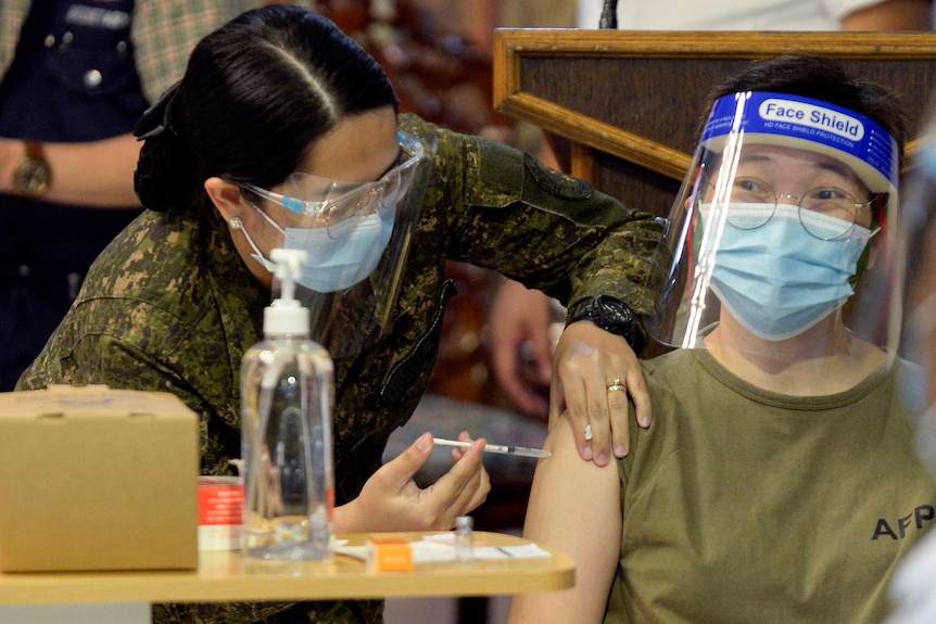 A woman in a facemask and wire rimmed glasses gets an injection from a nurse in her shoulder