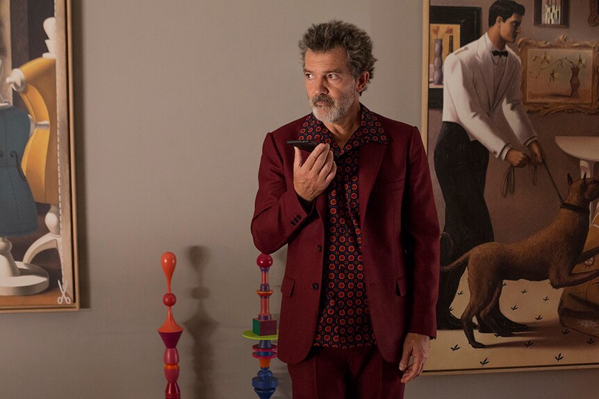 Antonio Banderas wears red and black patterned shirt with maroon suit and stands in gallery holding phone.
