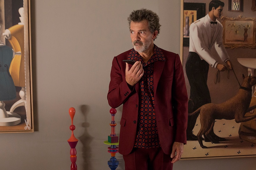 Antonio Banderas wears red and black patterned shirt with maroon suit and stands in gallery holding phone.